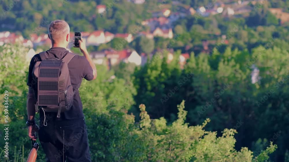 A young man in shorts, a T-shirt and a large backpack is traveling in Germany. A solar panel for charging a smartphone is hanging on the backpack. The man takes photos on a smartphone while hiking. A young man in shorts, a T-shirt and a large backpack is traveling in Germany. A solar panel for charging a smartphone is hanging on the backpack. The man takes photos on a smartphone while hiking.