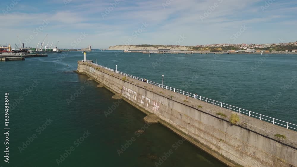 concrete pier with a lighthouse. The water is a deep blue and a city with cliffs are in the distance concrete pier with a lighthouse. The water is a deep blue and a city with cliffs are in the distance