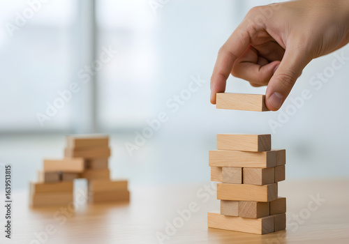 Building a business structure with wooden blocks. A hand carefully adds another level to a block tower, symbolizing strategic growth, project development, and building a strong foundation with precisi