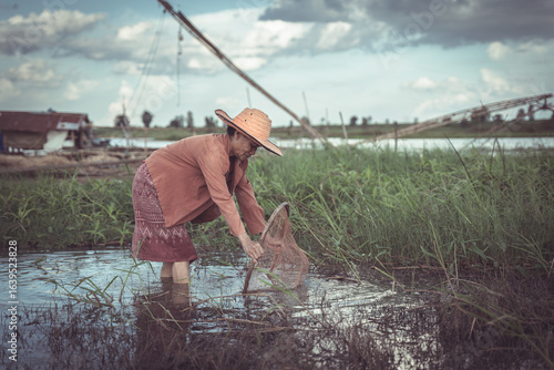 Woman in traditional clothing fishing with a net in shallow water surrounded by tall grass on a serene landscape with blue sky and clouds