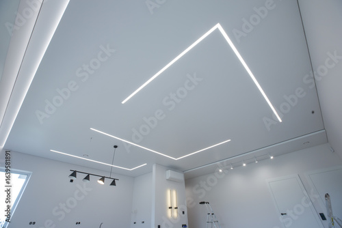 modern room interior with an emphasis on the ceiling. The ceiling is equipped with LED linear lights, creating a geometric pattern. Track lights, a pendant light and wall sconces are also visible.