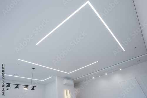 modern room interior with an emphasis on the ceiling. The ceiling is equipped with LED linear lights, creating a geometric pattern. Track lights, a pendant light and wall sconces are also visible.