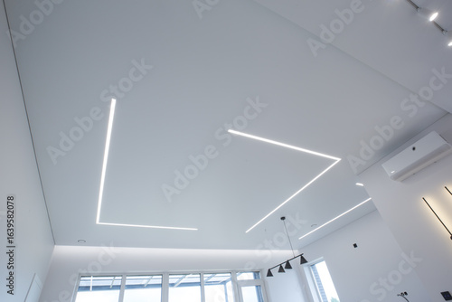 modern room interior with an emphasis on the ceiling. The ceiling is equipped with LED linear lights, creating a geometric pattern. Track lights, a pendant light and wall sconces are also visible.