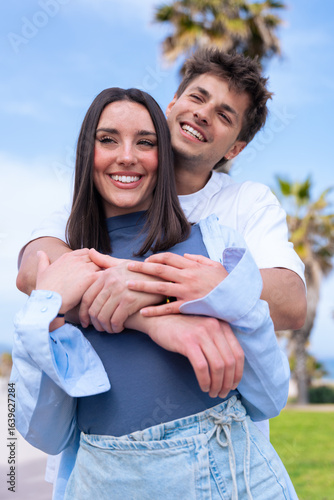 Vertical portrait Young couple embrace on a palm lined beach promenade, boyfriend laughing as girlfriend wraps arms around his shoulders from behind, capturing joyful love, travel vibes.