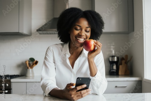 Modern beautiful African American female with smartphone in hand sitting on kitchen counter looking away at home and eating apple