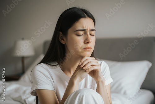 Worried young woman with eyes closed sitting near bed at home