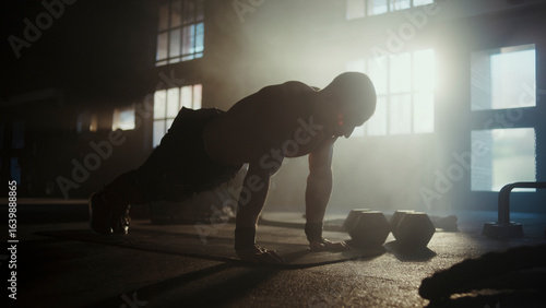Muscular athlete doing push-ups in a dramatic gym setting with backlight