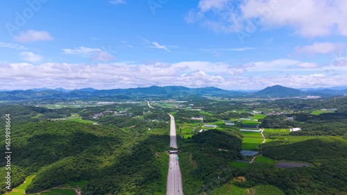 Aerial Drone View of Highway and Cars Driving Through Green Fields and Forest, with Vast Summer Landscape, Blue Sky, and Clouds for a Healing Scenery