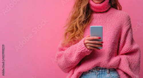 Woman in cozy pink sweater holds smartphone against pink background, showcasing casual and stylish look. Her long hair and painted nails add to vibrant and modern aesthetic