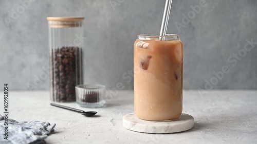 Coffee latte with milk and ice in a glass is stirred with a straw on a marble stand on a light background with coffee beans and morning shadows. The concept of a summer refreshing craft drink.