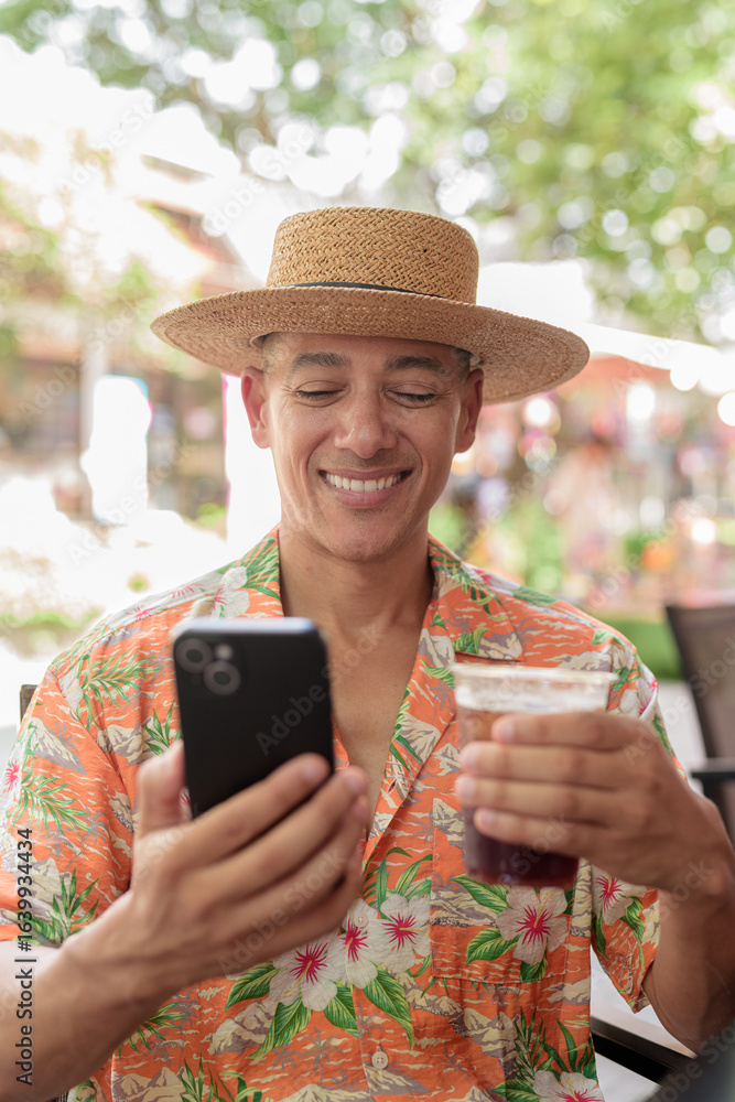 Fototapeta premium Hispanic man holding iced coffee and using phone at cafe wearing Panama hat and Hawaiian shirt