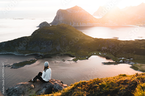 Young woman hiker standing on edge of cliff and watching sunrise or sunset in Lofoten Islands. Midnight sun over Lofoten Islands
