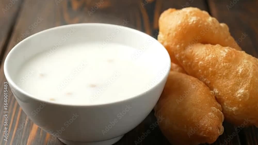 Bowl of white liquid with grains and fried dough sticks sit on a dark wooden surface Bowl of white liquid with grains and fried dough sticks sit on a dark wooden surface