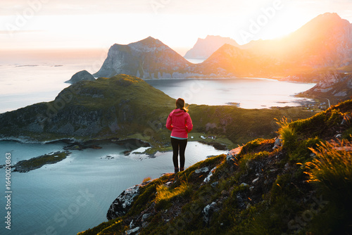 Young woman hiker standing on a rocky cliff in Lofoten Islands, Norway, during sunrise in summer, with the golden midnight sun lighting the fjords.