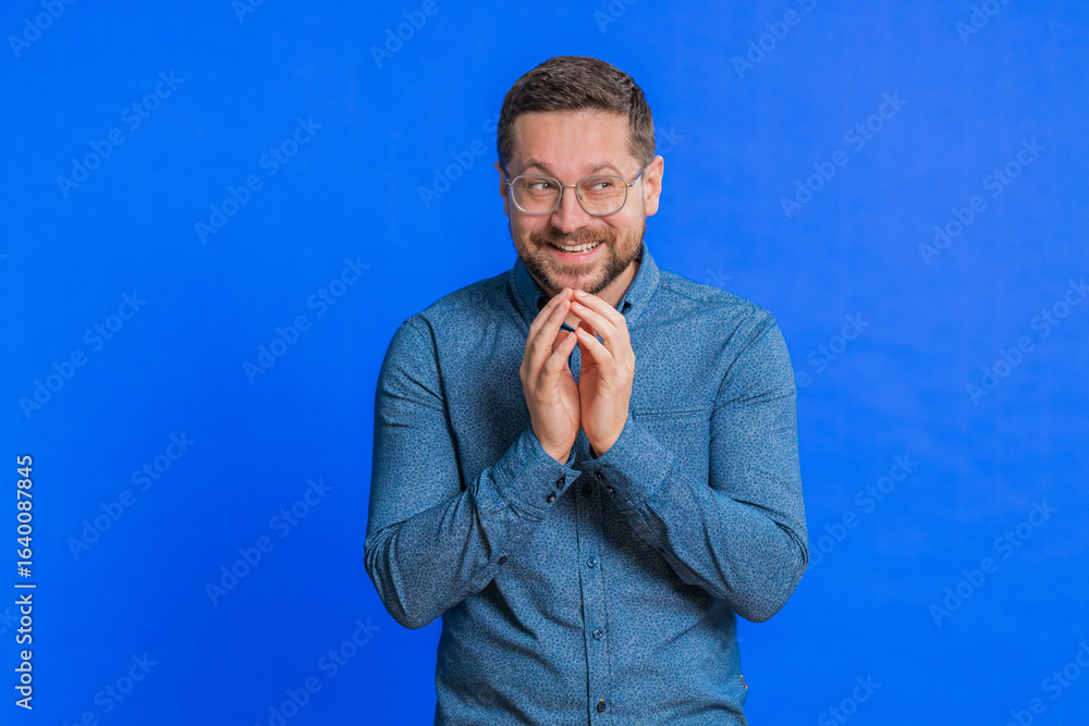 Fototapeta premium Sneaky cunning Caucasian middle-aged man in glasses with tricky face gesticulating and scheming evil plan, thinking over devious villain idea, cheats, jokes, pranks. Guy isolated on blue background