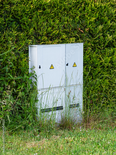 Outdoor electrical cabinet with high voltage warning signs surrounded by tall grass and green bushes in summer.