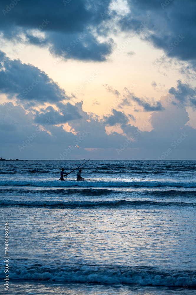 Fototapeta premium Two fishermen silhouetted at dusk cast their nets in the Arabian Sea from Siridao Beach, Panjim, Goa, beneath a monsoon sky painted orange with clouds over the calm evening waves.