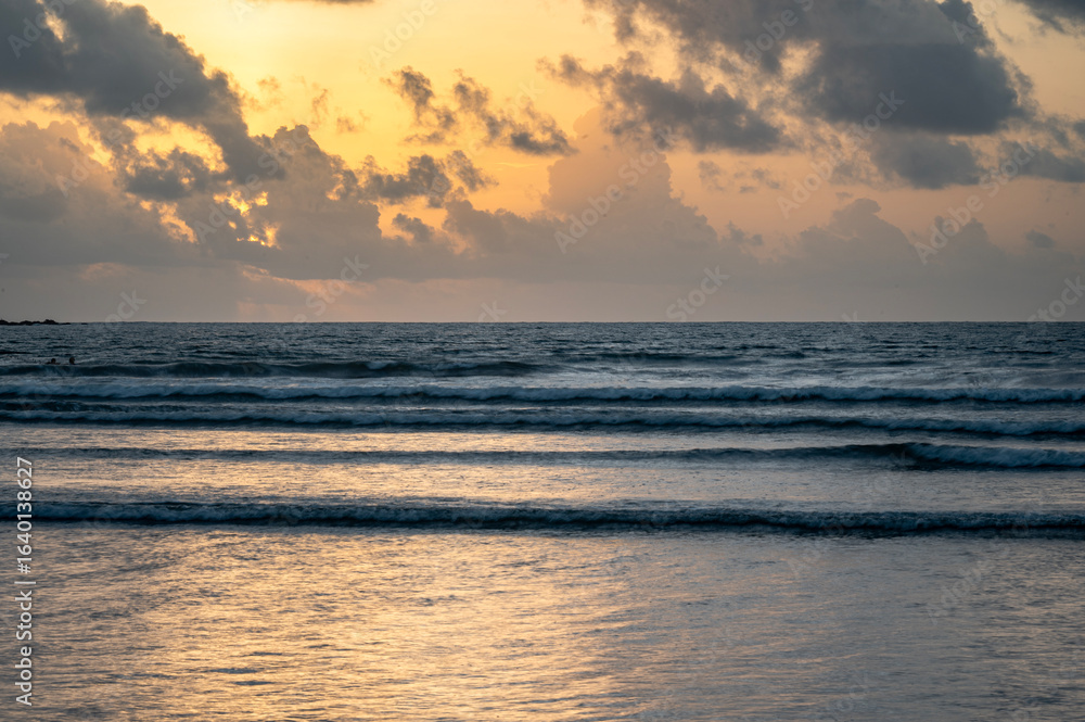 Obraz premium Sunset view of Vagator Beach Goa with Arabian Sea, golden yellow sky, dramatic grey monsoon clouds, and shimmering water reflecting the sun’s warm light in the evening.
