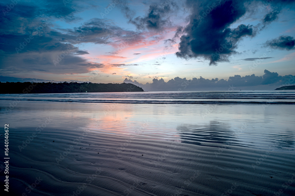 Fototapeta premium Landscape shot of Miramar Beach, Goa, India, after sunset at dusk with wet sand line textures, waves, two backlit cliffs, cloud line at horizon, and surreal sky with yellow, pink, orange, and dramatic