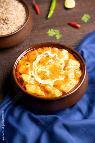 фотографія A top shot of Paneer Butter Masala, cottage cheese slow-cooked in rich buttery tomato and cashew nut gravy, served in a rustic brown pot on blue satin with green and red chilies