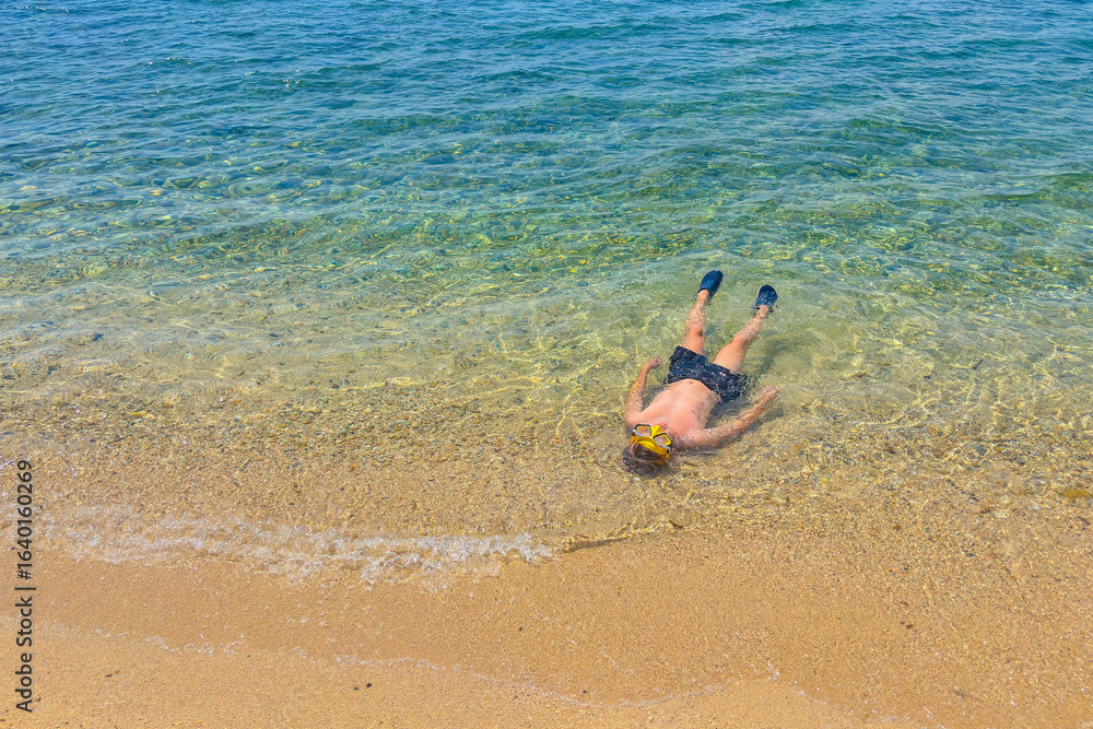 Obraz premium young boy floating on his back in the shallow, clear turquoise sea with sand on a sunny day, top view of a child wearing a yellow snorkel mask and flippers, concept of summer vacation, family holidays