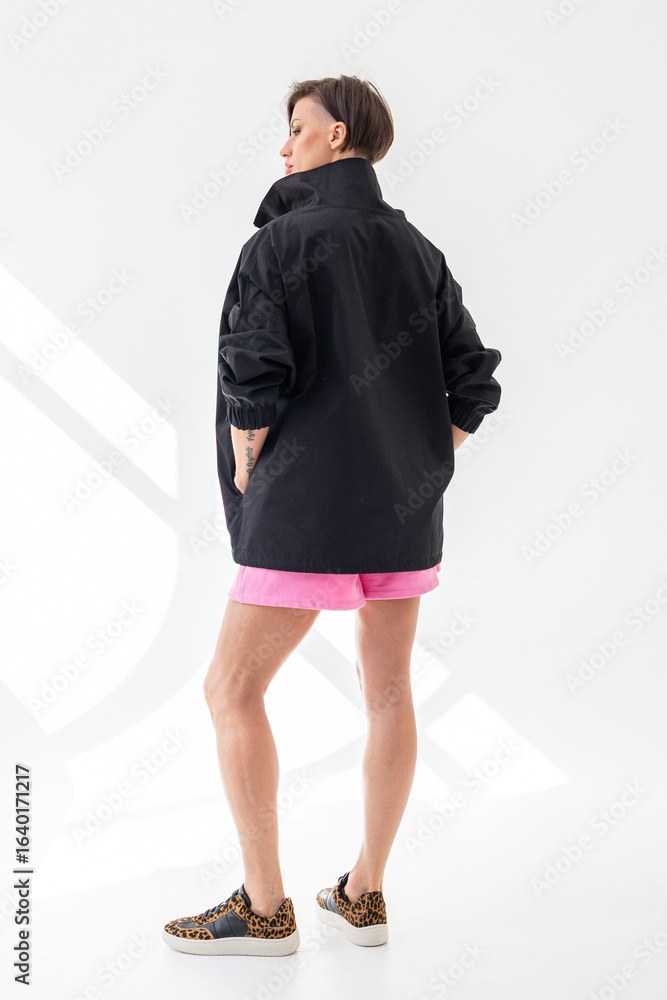 Fototapeta premium Elegant woman in black windbreaker jacket and pink shorts on light background. Fashionable female style. Studio photography. Vertical photo. High quality photo