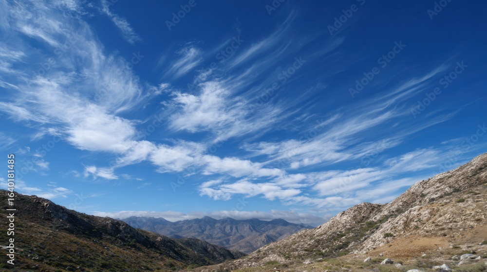 Under a bright blue sky, wispy clouds drift above rugged mountain peaks. The landscape features rolling hills, creating a serene outdoor atmosphere perfect for exploration.