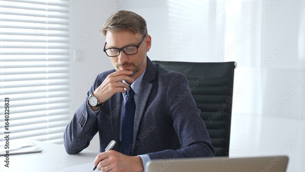 Adult businessman wearing blue suit and tail is concentrating and thinking while working at the desk in a bright, modern office, demonstrating professionalism and efficiency. Business people concept Adult businessman wearing blue suit and tail is concentrating and thinking while working at the desk in a bright, modern office, demonstrating professionalism and efficiency. Business people concept