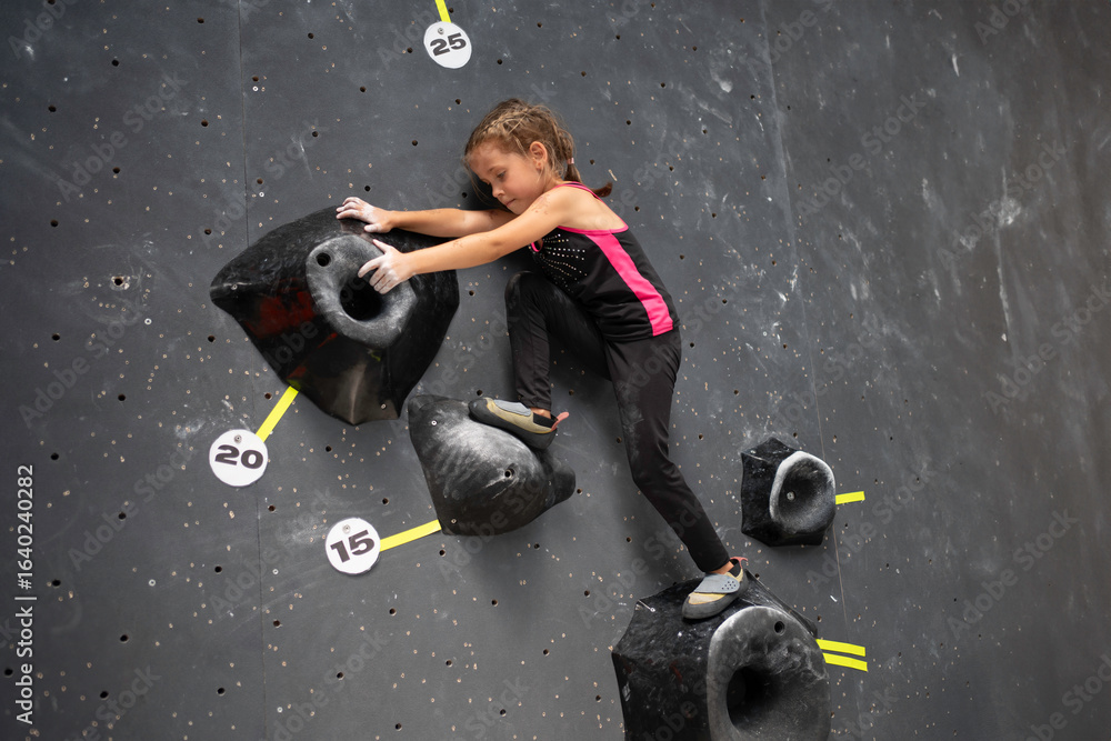 Fototapeta premium Determined girl climbing artificial wall at bouldering center. Brave and strong female child tackling mock wall, full body side view of sporty girl in action