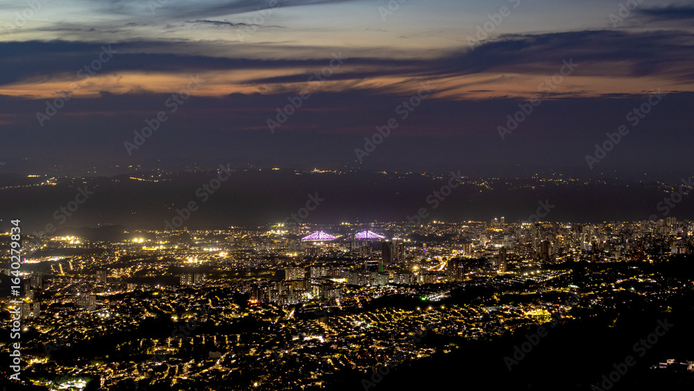 Fototapeta premium Night view of Bucaramanga, Colombia, with a glowing city skyline, bright stadium lights, and a twilight sky fading into darkness, captured from an elevated viewpoint.