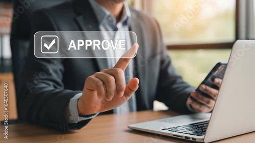 Man using laptop and phone with approve button on screen in office setting for business approval task