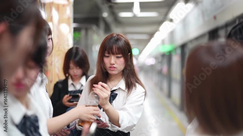 Slow-motion. Japanese high school girls in uniform wait on a station platform, talking while using smartphones and checking a timetable as rails gleam. Shanghai's Pudong New District. China. Spring.