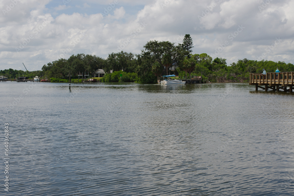 Fototapeta premium Serene Waterfront Scene with Dock, Boat, and Lush Greenery people fishing. Calm mangrove area featuring a dock, a boat, and green vegetation under a partly cloudy sky, conveying tranquility. 