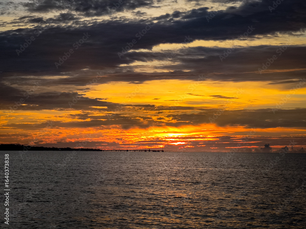 Fototapeta premium Vivid sunset with intense orange and yellow hues over the ocean, featuring a distant pier silhouette beneath dramatic layered clouds.