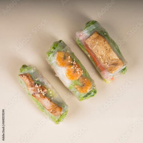 Photography Three fresh spring rolls with various fillings grilled chicken, shrimp, and tofu wrapped in rice paper with lettuce and sprinkled with sesame seeds, arranged on a light beige background