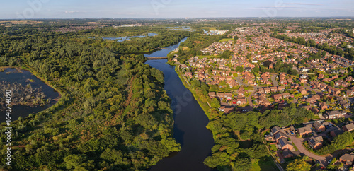 Aerial panoramic view of Woolston, near Warrington, with the River Mersey winding past residential areas, dense woodlands, and a wetlands