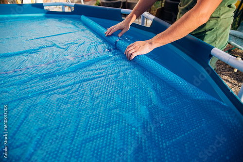 The blue pool cover is assembled into a roll by men's hands, the concept of summer recreation and pool maintenance.
