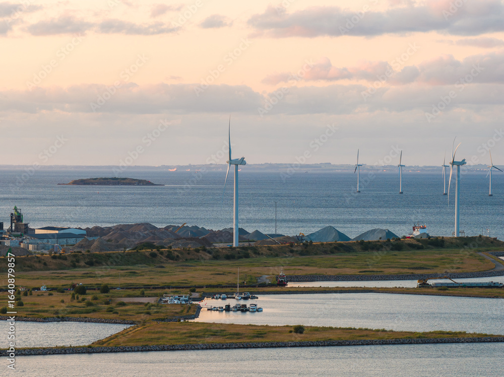 Obraz premium Aerial view of Copenhagen's coastal area with wind turbines near the water, industrial structures, greenery, and a serene sea in the background.