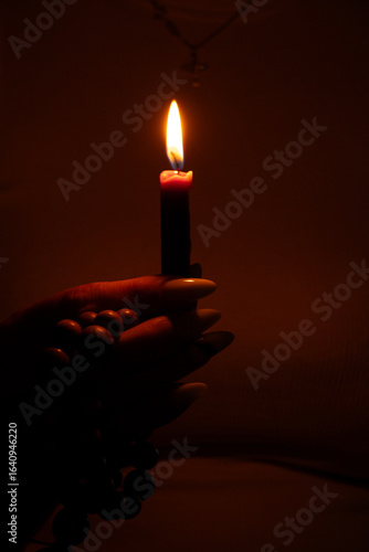 Woman's hand holding a lit candle and wooden rosary beads in the dark at home, flame illuminates fingers, faith