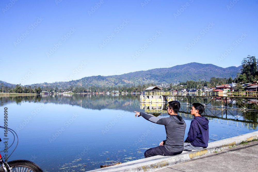 Fototapeta premium Rear View Of Two Male Quietly Sit On A Lakeside Edge, Enjoying The Peaceful Water View