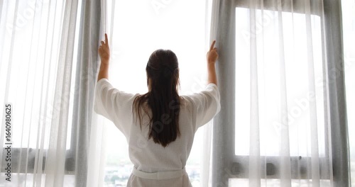 A beautiful woman in a bathrobe, opening the curtains to greet the morning sunlight.Urban living lifestyle in bed room 