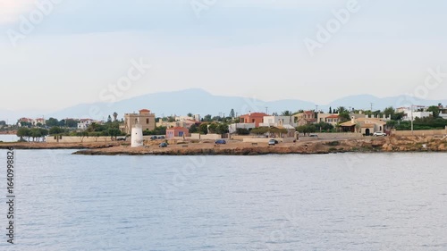 Wallpaper Mural Tranquil coastal scene on Aegina Island Greece with calm sea, rocky pier, historic white lighthouse, pastel houses, trees, and distant mountains under a light cloudy sky, evening time Torontodigital.ca