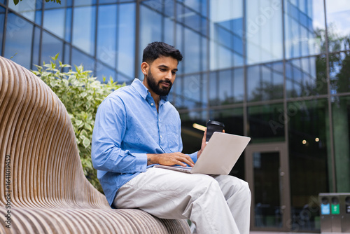 A man sits on a bench, using a laptop outdoors, with a coffee cup in hand, in front of a modern building.