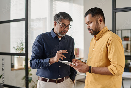 Medium shot of two Indian men standing in office looking at digital tablet screen discussing business plan