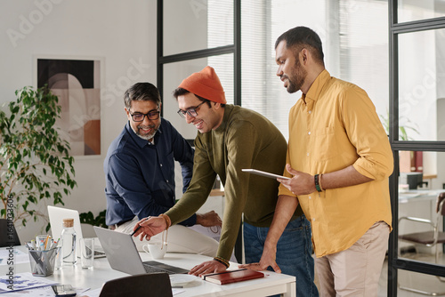 Medium long shot of three cheerful young and mature Indian men working on business project in modern office