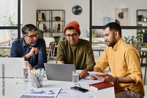 Group of three Indian men sitting at table in modern office working on business project, looking at laptop screen and discussing plan