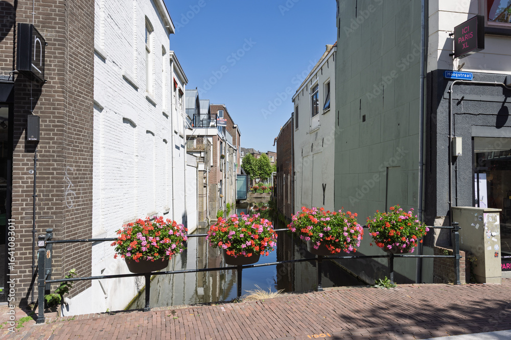 Naklejka premium A charming canal view framed by historic buildings, with vibrant flower baskets adorning the railing of a bridge over the tranquil water. Gouda, Netherlands, 29 June 2025.