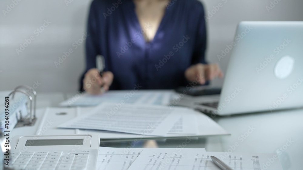 Fototapeta premium Magnifying glass, white calculator, pen and documents are lying on accountant's desk. Businesswoman wearing blue dress is working with documents on the background. Business and audit concept