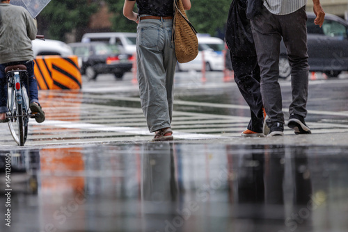 Urban rain scene. Women dressed in faded jeans with big woven straw shoulder bag is focus of pedestrians waiting in rain at busy zebra crossing. Wet pavement and reflects surrounding elements.
