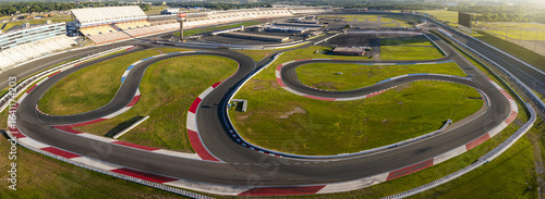 Concord, United States - 28 July 2025: Aerial view of a race track's winding asphalt curves cutting through vibrant green fields, contrasted by the stark white and red safety barriers.
